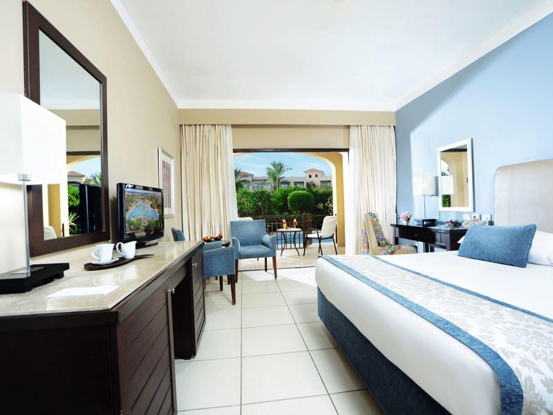 Lichte hotelkamer met kingsize bed, tv, bureau, stoelen en balkon met uitzicht op tuin.