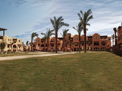 Eine Hotelanlage mit Rasenfläche, Palmen und zweistöckigen Gebäuden unter einem bewölkten Himmel.