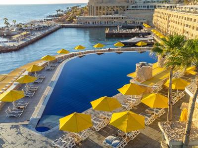 Großer Hotelpool mit gelben Sonnenschirmen und Blick auf das Meer bei Sonnenuntergang.