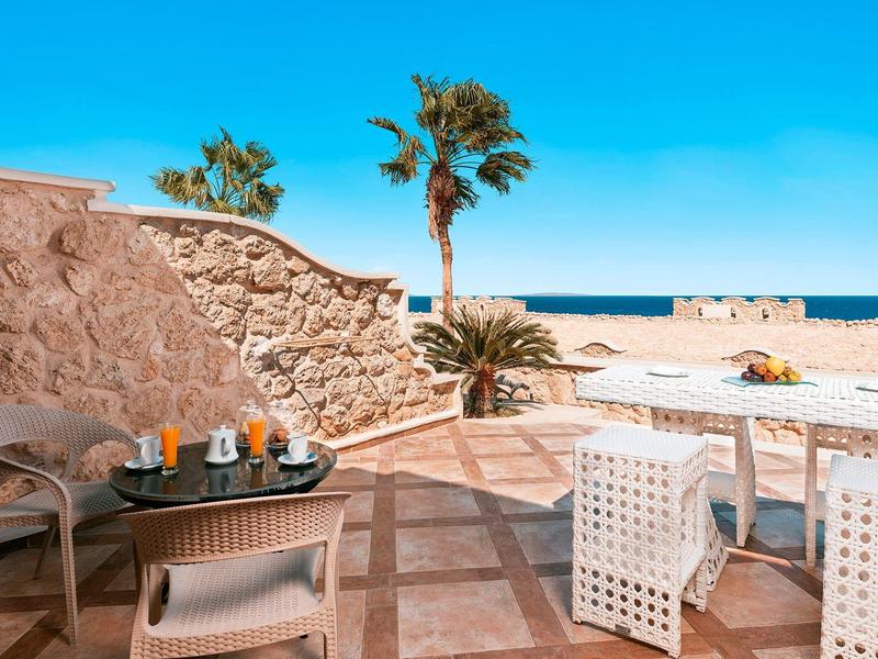 Buitenzitgedeelte met stoelen en tafels met uitzicht op een strand en heldere blauwe lucht.