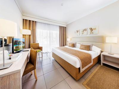 Habitación de hotel luminosa con cama doble, escritorio, silla y grandes ventanas con cortinas.