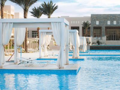 Schwimmbad mit weißen Baldachin-Betten und Palmen vor einem Hotelgebäude