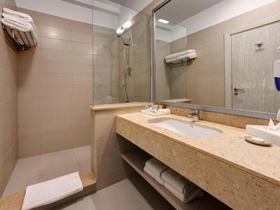 Baño de hotel luminoso con ducha, espejo grande y encimera de mármol beige.