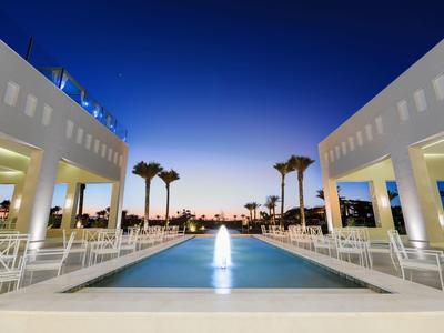 Piscina moderna con juegos de agua entre edificios blancos al atardecer.