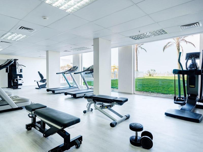 Gimnasio moderno con pesas, cintas de correr y grandes ventanas con vista a área verde.