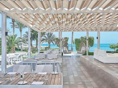 Restaurante al aire libre con techo de madera, mesas y vista a palmeras y al mar.