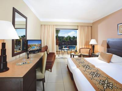 Bright hotel room with double bed, desk, and balcony view of pool.