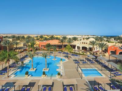 Large hotel pool with many sun loungers, surrounded by palm trees and buildings under a blue sky.