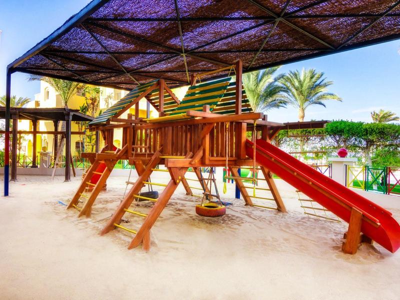 Área de juegos cubierta con estructura de madera y tobogán rojo en un hotel o resort.