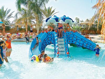 Kinder spielen im flachen Pool mit blauem Delfin-Wasserrutschen-Spielplatz unter Palmen.