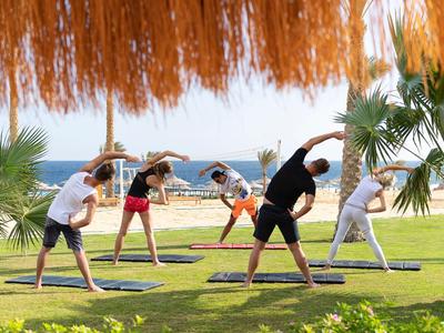 Personas hacen gimnasia sobre esterillas de yoga al aire libre en la playa bajo un techo de paja.