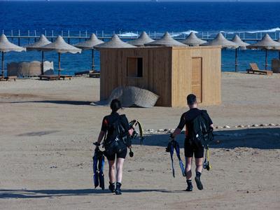 Two people with diving gear walk on a sandy beach toward the sea with sun umbrellas.