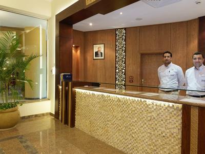 Hotel reception area with wooden counter, two staff members, and indoor plants.