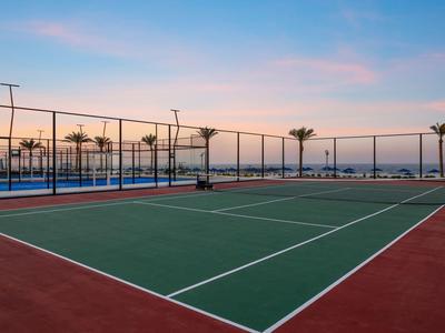 Leeg tennisveld met omheining en palmbomen bij zonsondergang aan zee.