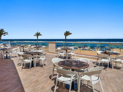 Terrasse d'hôtel en bord de mer avec tables rondes, chaises blanches et palmiers sous un ciel clair.