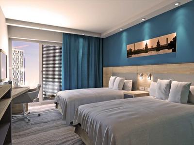 Modernes Hotelzimmer mit zwei großen Betten, blau-grauen Wänden, Schreibtisch und Stadtbild-Bild.