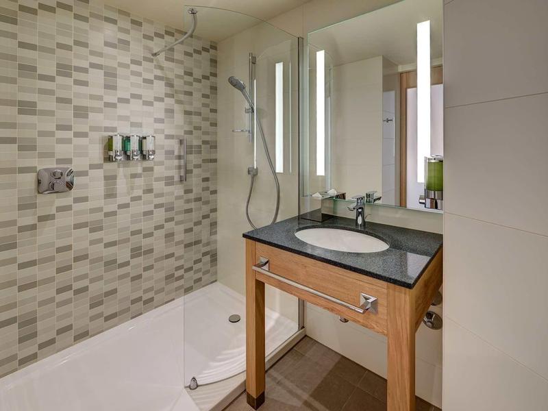 Moderne Dusche mit Mosaikwand, Holzmöbel und beleuchtetem Spiegel in hellem Badezimmer.