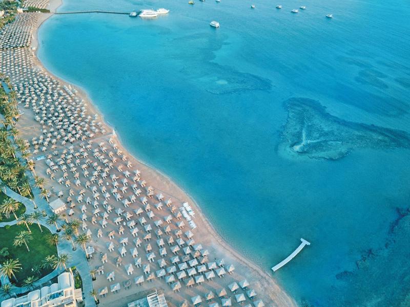 Luftaufnahme eines langen, sandigen Strandes mit vielen Sonnenschirmen und klar blauem Meer.