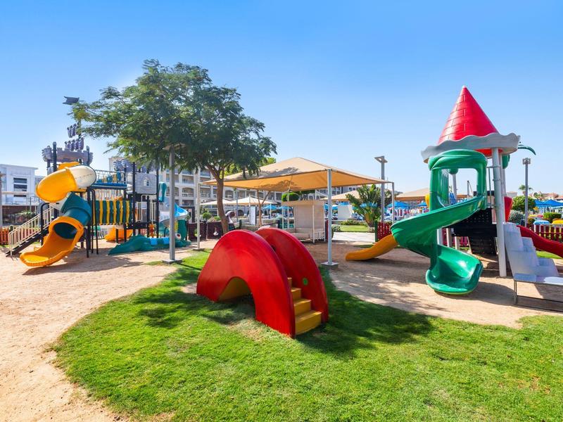 Kinderspielplatz mit bunten Rutschen, Klettergeräten, Sandflächen und schattigen Bäumen bei blauem Himmel.