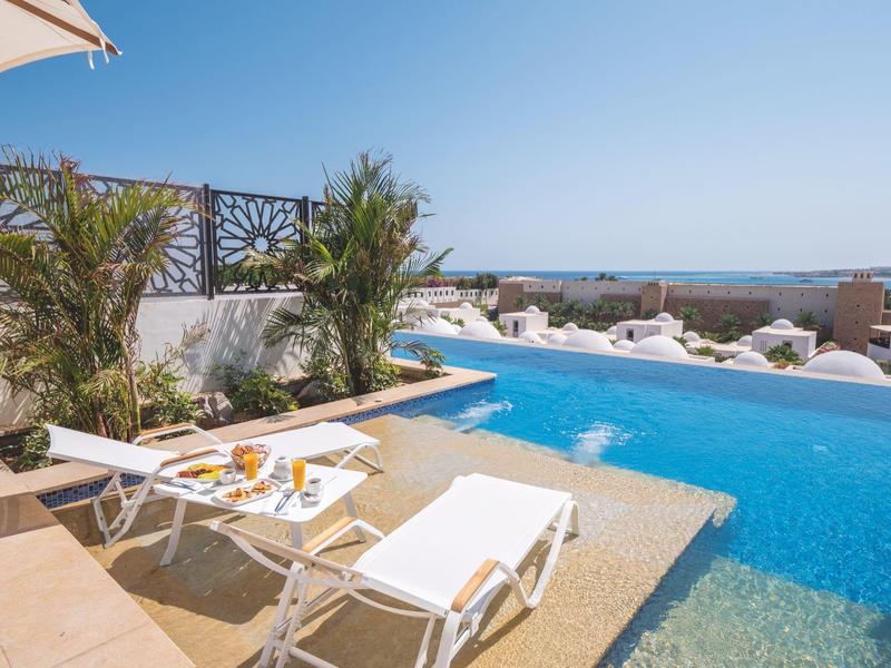 Poolbereich mit Liegestühlen, Pflanzendekoration und Meerblick bei klarem Himmel.