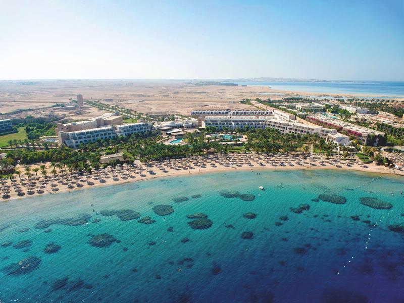 Luftaufnahme eines Resorts am Meer mit Strand, Liegestühlen und klarem blauem Wasser.