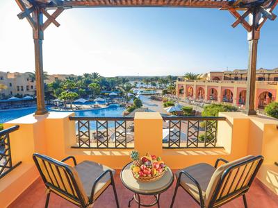 Balcony with two chairs and table with fruit bowl, view of hotel pool and buildings in sunlight.