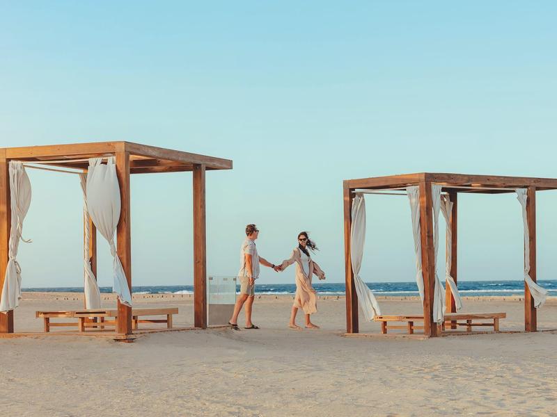 Zwei Personen halten Händchen und laufen zwischen zwei Holzpavillons am Strand.