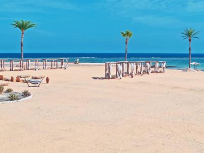 Weite Sandfläche mit Palmen, Sonnenliegen und blauem Meer im Hintergrund unter klarem Himmel.