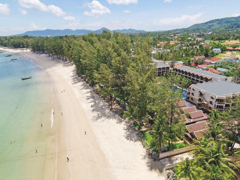 Aerial view of a long sandy beach with clear water, trees, and hotels in the background.