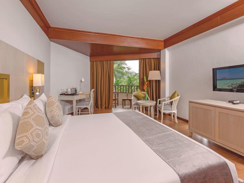 Spacious hotel room with large bed, desk, chairs, and balcony overlooking greenery.