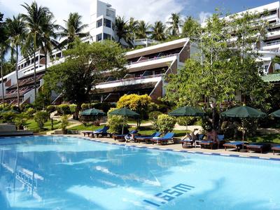 Ein großer Pool mit klarem Wasser vor einem Hotel, umgeben von Palmen und grünen Pflanzen.