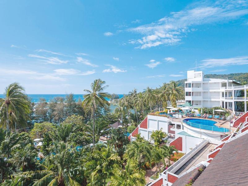 Hotel met zwembad, palmbomen en zeezicht onder blauwe lucht.