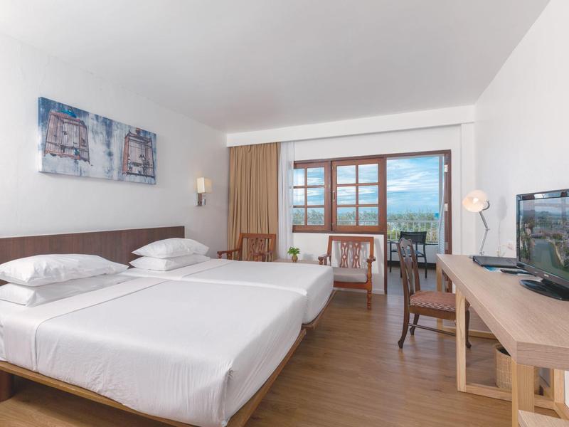 Lichte hotelkamer met twee eenpersoonsbedden, bureau, tv en balkonzicht.