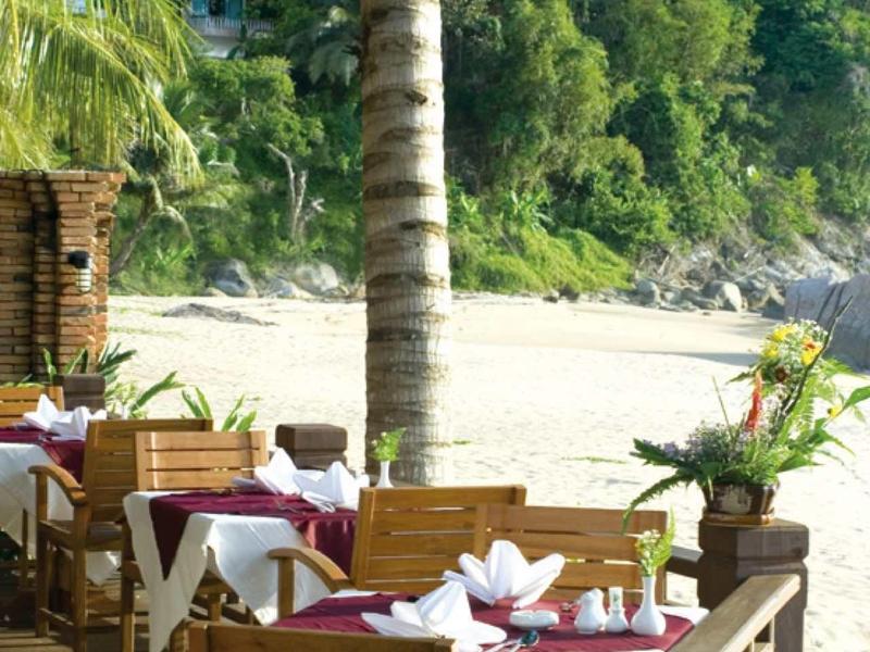Holzterrasse mit gedeckten Tischen am Strand, Palmen und dichter grüner Vegetation im Hintergrund.