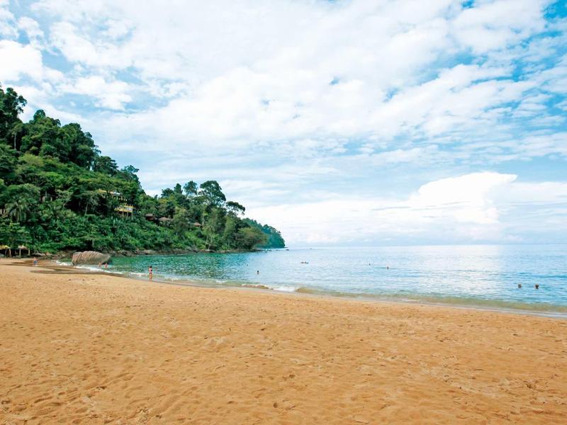 Breiter Sandstrand mit grün bewaldetem Felsen und ruhigem blauem Meer unter bewölktem Himmel.