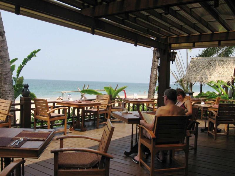 Offene Strandbar mit Holzmöbeln, Palmen und Blick auf das Meer bei klarem Himmel.