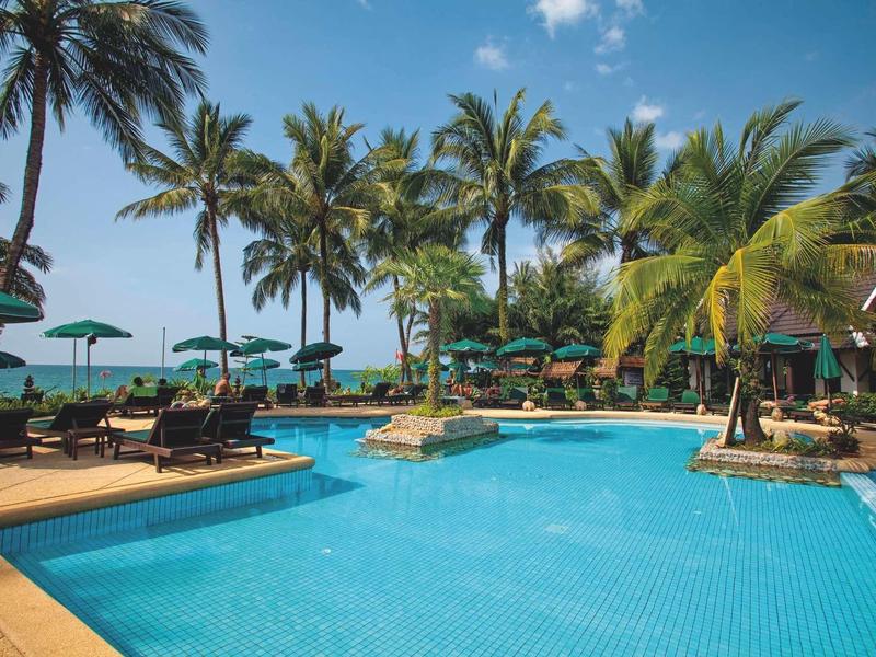 Großer Außenpool mit blauen Fliesen, umgeben von Palmen, Liegestühlen und Sonnenschirmen bei blauem Himmel.
