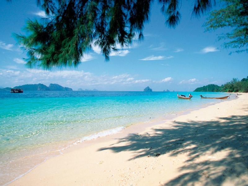 Strand mit weißem Sand, türkisblauem Wasser, grünen Bäumen und blauem Himmel mit Wolken.