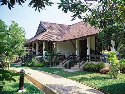 Ein Haus mit braunem Ziegeldach, Veranda und grünem Garten mit Weg und Bäumen im Vordergrund.