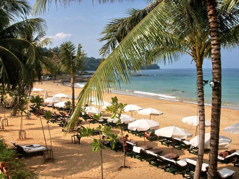 Strand mit Palmen, Sonnenliegen und weißen Sonnenschirmen am Meer unter blauem Himmel.
