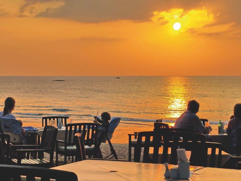 Menschen sitzen an Tischen am Strand und beobachten den orangefarbenen Sonnenuntergang über dem Meer.