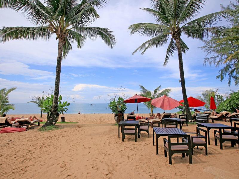 Strand mit Sand, Palmen, Stühlen, roten Sonnenschirmen und Blick auf ruhiges Meer.