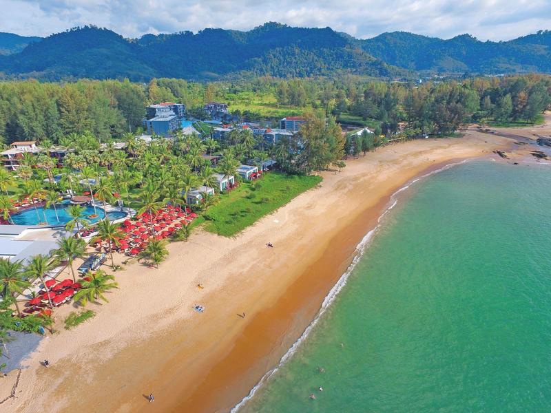 Langer Sandstrand mit klarem grünem Wasser, angrenzend an grüne Hügel und Gebäude im Hintergrund.