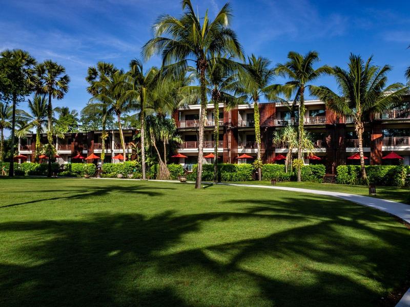 Hotel mit roten Fassaden umgeben von grünen Palmen und Rasen unter blauem Himmel.