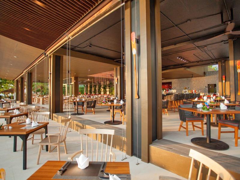 Moderner Außenbereich eines Restaurants mit Holztischen, weißen Stühlen und großer Glasfront.