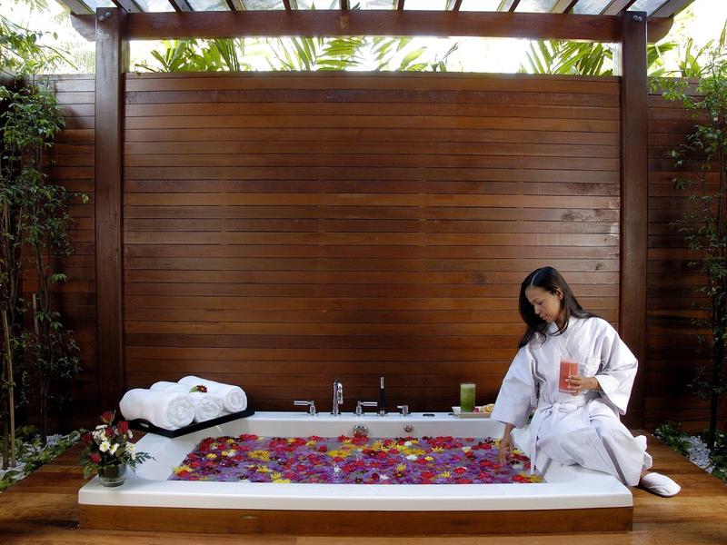 Frau in weißem Bademantel am gefüllten Bad mit Rosenblättern, Holzwand und Pflanzen im Hintergrund.