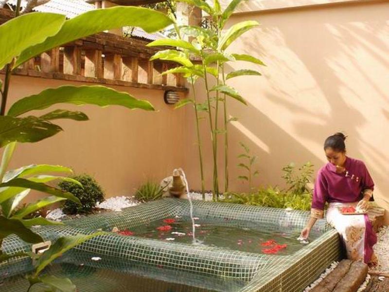 Een klein rechthoekig zwembad met drijvende bloemen, omgeven door planten en een persoon die op een bankje zit.