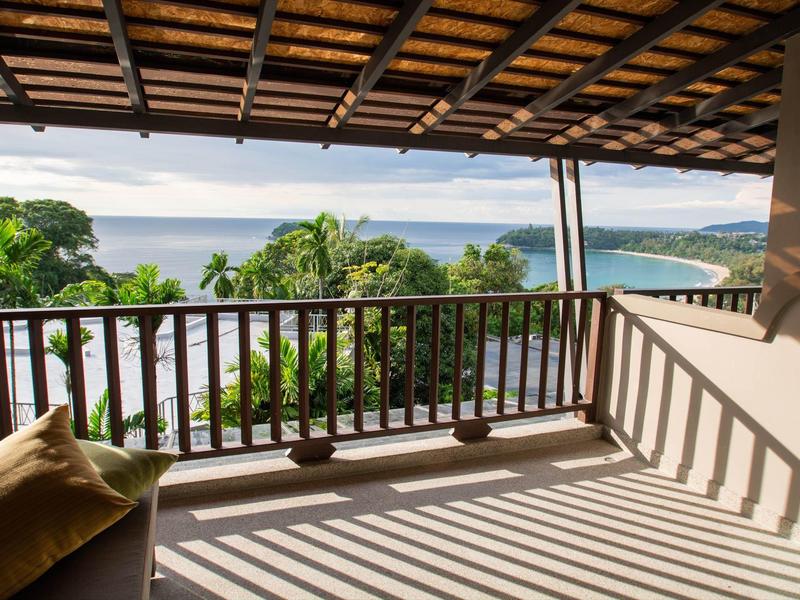 Balkon met houten reling met uitzicht op groene bomen en de zee onder een bewolkte lucht.