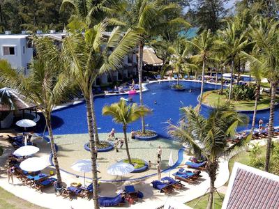 Großer, ovaler Pool umgeben von Palmen, Sonnenschirmen und Liegestühlen in einer tropischen Anlage.