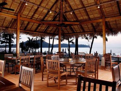 Offenes Restaurant mit Holztischen und -stühlen, Blick auf Palmen und Meer bei Sonnenuntergang.